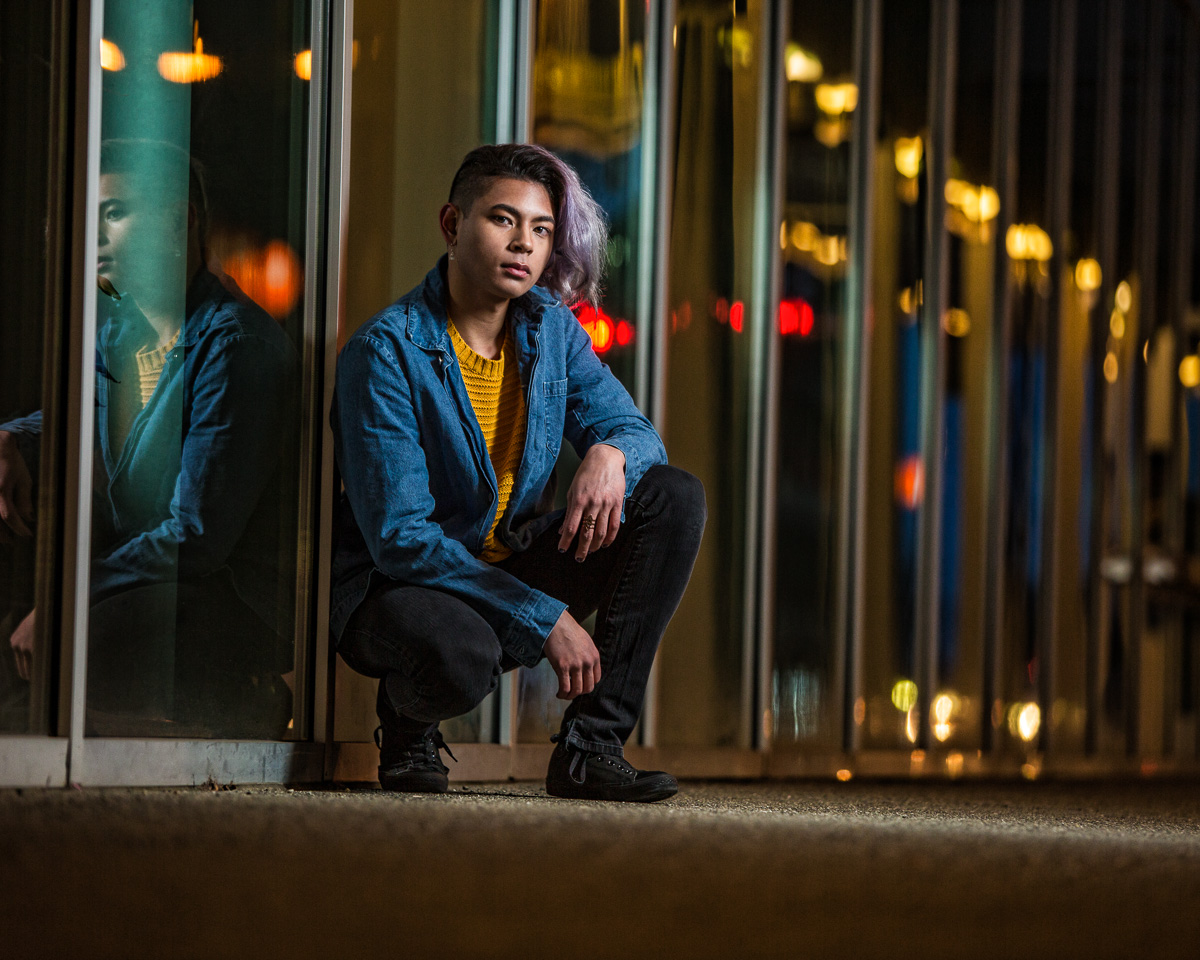 High school senior boy kneeling down by some windows, with streetlights reflecting