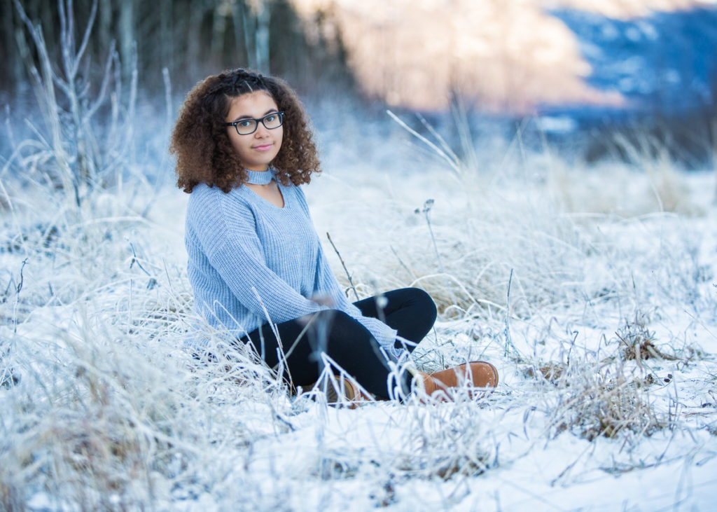 High school senior girl sitting in the snow in a blue long sleeve shirt