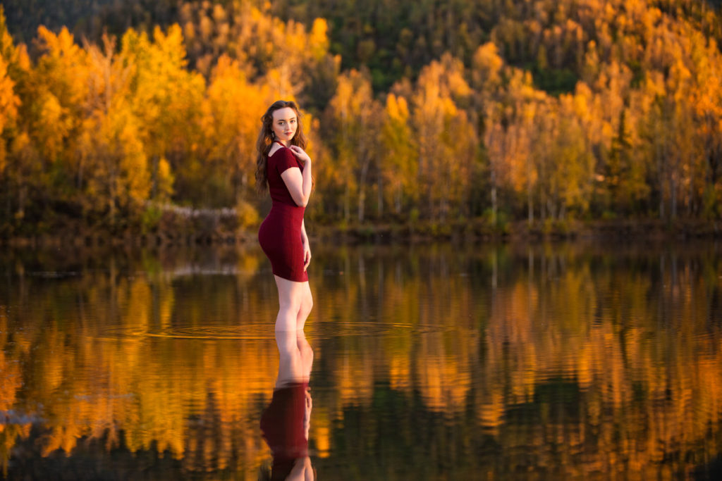 High school senior girl standing in a lake wearing a red dress in front of fall trees