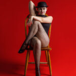 Hilary Hanson-1896 girl sitting with her legs crossed on and chair on a red background