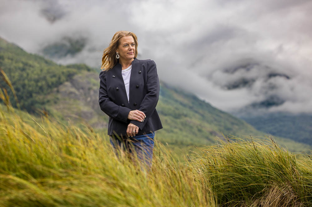 Mature woman standing in a windy grass field with mountains in the background