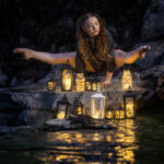 Kira Moore-7855 girl on her hands on rocks with lanterns in front of her with water reflecting