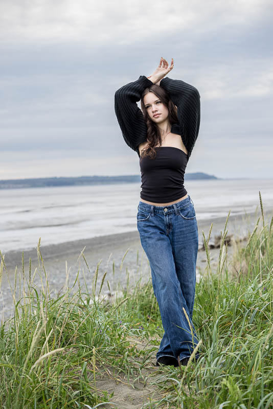 girl wearing a black sweater and blue jeans standing in the grass with ocean side behind her