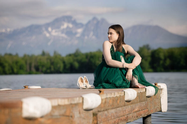 senior photos of a girl in a green dress sitting on a lake dock with shoes beside her