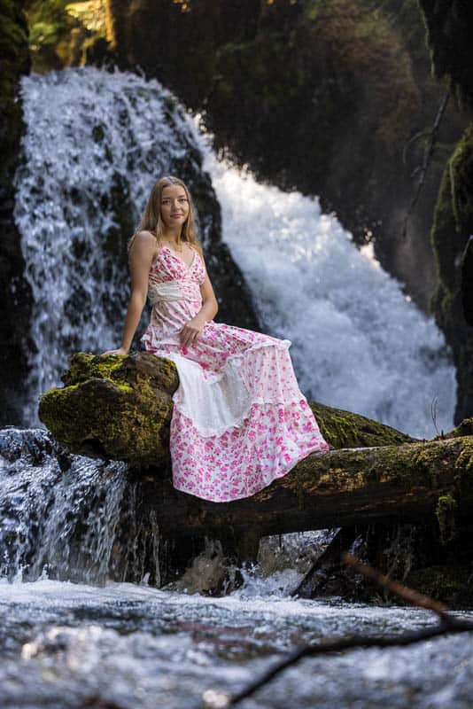 girl in a dress sitting on a log over water with a waterfall behind her