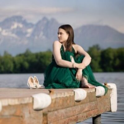 Long haired high school senior girl in a green dress sitting on a dock with a lake and mountains in the background