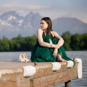 Long haired high school senior girl in a green dress sitting on a dock with a lake and mountains in the background