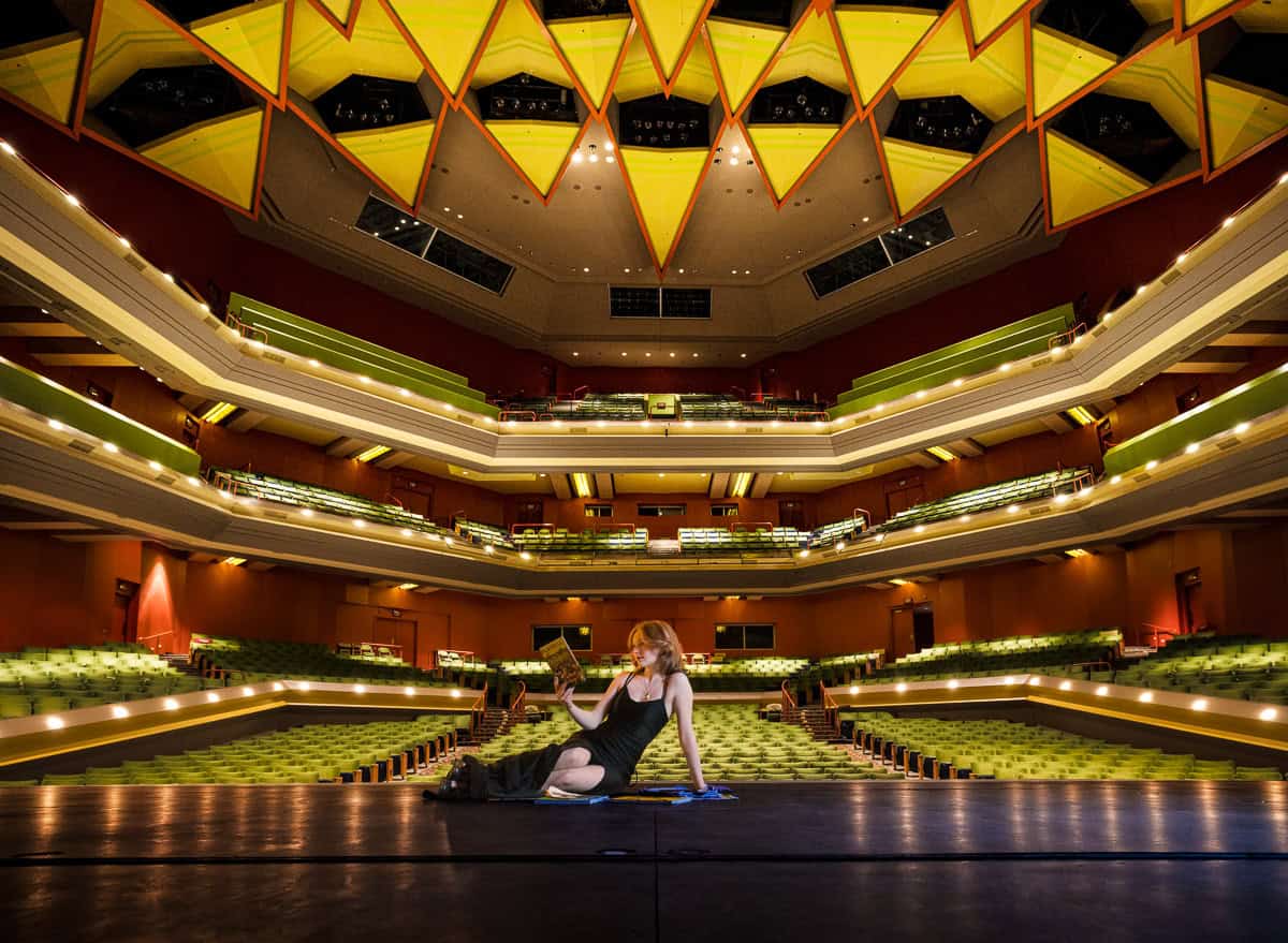 High school senior girl in a long formal dress reading a program sitting a stage with theater seating in the background