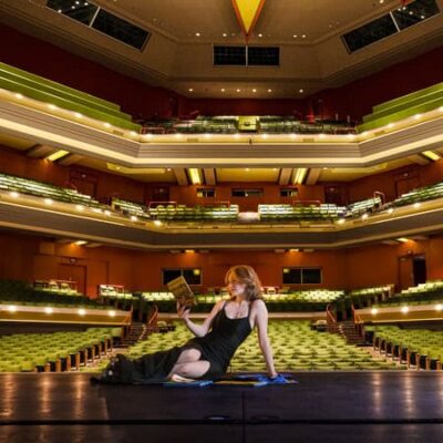 High school senior girl in a long formal dress reading a program sitting a stage with theater seating in the background