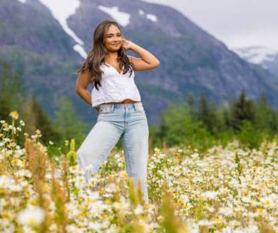 High school senior girl wearing denim pants and a white short sleeved top standing in a field of flowers with mountains in the background