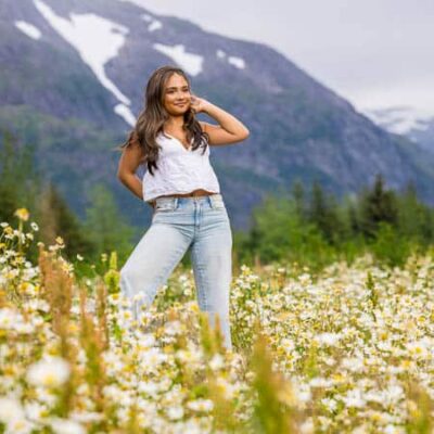 High school senior girl wearing denim pants and a white short sleeved top standing in a field of flowers with mountains in the background